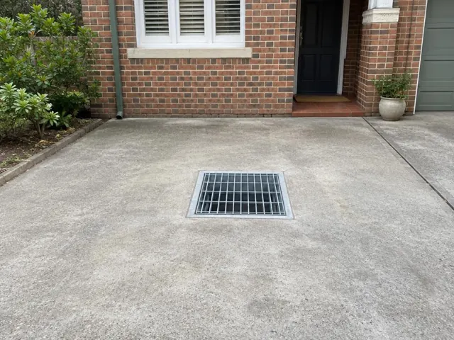 Concrete driveway with a metal drain grate outside a brick house in Turramurra