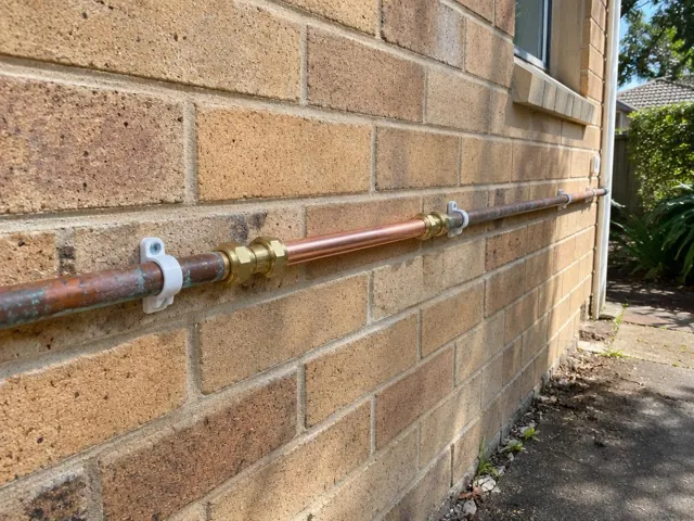 Repaired copper water pipe running along a brick exterior wall beside a side path in Turramurra