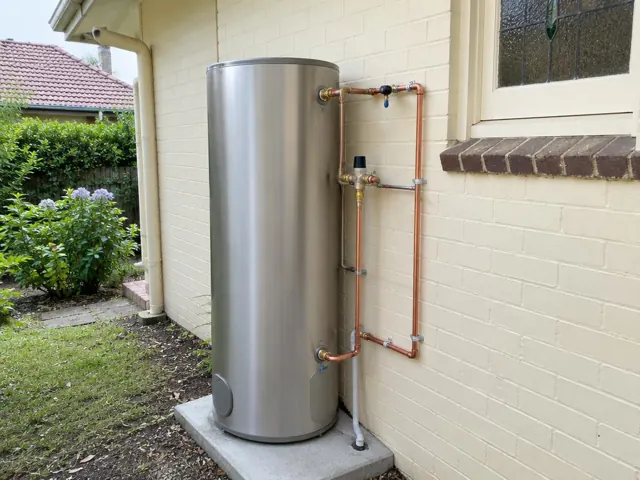 Outdoor stainless steel hot water cylinder beside a cream brick house wall and window in Wahroonga